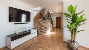 A modern living room with a flat-screen TV, a white console table, a staircase with a stone wall, and a large green plant. - Sunnyside Villa with private pool - 10 min from the beach! (Calheta)