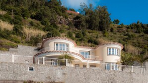 A two-story house with a curved roofline, stone walls, and a balcony with a railing. - Sunnyside Villa with private pool - 10 min from the beach! (Calheta)