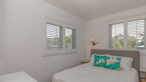 Un dormitorio con una cama, dos ventanas con persianas, una mesita de noche y una lámpara.
