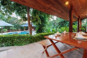 Un área de comedor al aire libre con una mesa de madera dispuesta para dos, con vistas a una piscina y abundante vegetación.