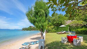 A beach with clear blue water, a sandy shore, and a grassy area with lounge chairs and a barbecue. - TED Villas (Koh Samui)