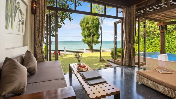 A modern room with a large window overlooking a beach and ocean. There is a sofa, a table with a laptop, and a wicker chair. - TED Villas (Koh Samui)