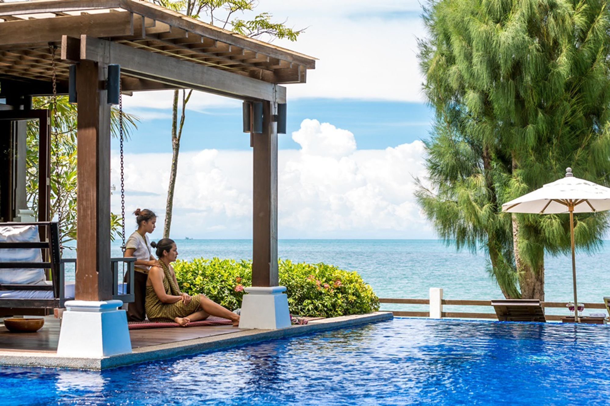 Un espace aménagé au bord de la piscine, avec une pergola en bois, une table de massage et un(e) massothérapeute qui prodigue un soin à un(e) client(e).