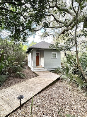 Une petite maison de plain-pied avec une porte rouge et une terrasse en bois menant à l’entrée.