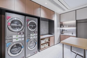 A modern laundry room with four stacked washing machines, a small table, and a shelf with toiletries. - JI Hotel (Shanghai)