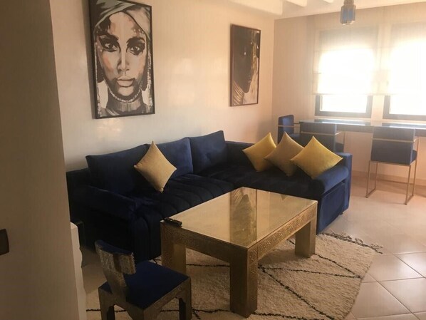 A living room with a dark blue sofa, a glass coffee table, and a patterned rug. There are two paintings on the wall and a window providing natural light.