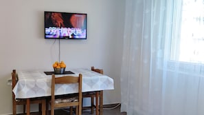 Ruokailutila, jossa on kahdelle katettu pöytä, seinälle asennettu televisio ja ikkuna, jossa on läpikuultavat verhot.