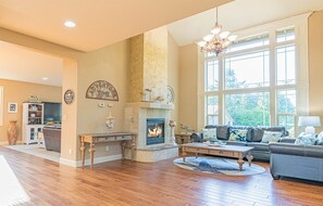 A spacious living room with hardwood flooring, a fireplace, a large window, and a chandelier.