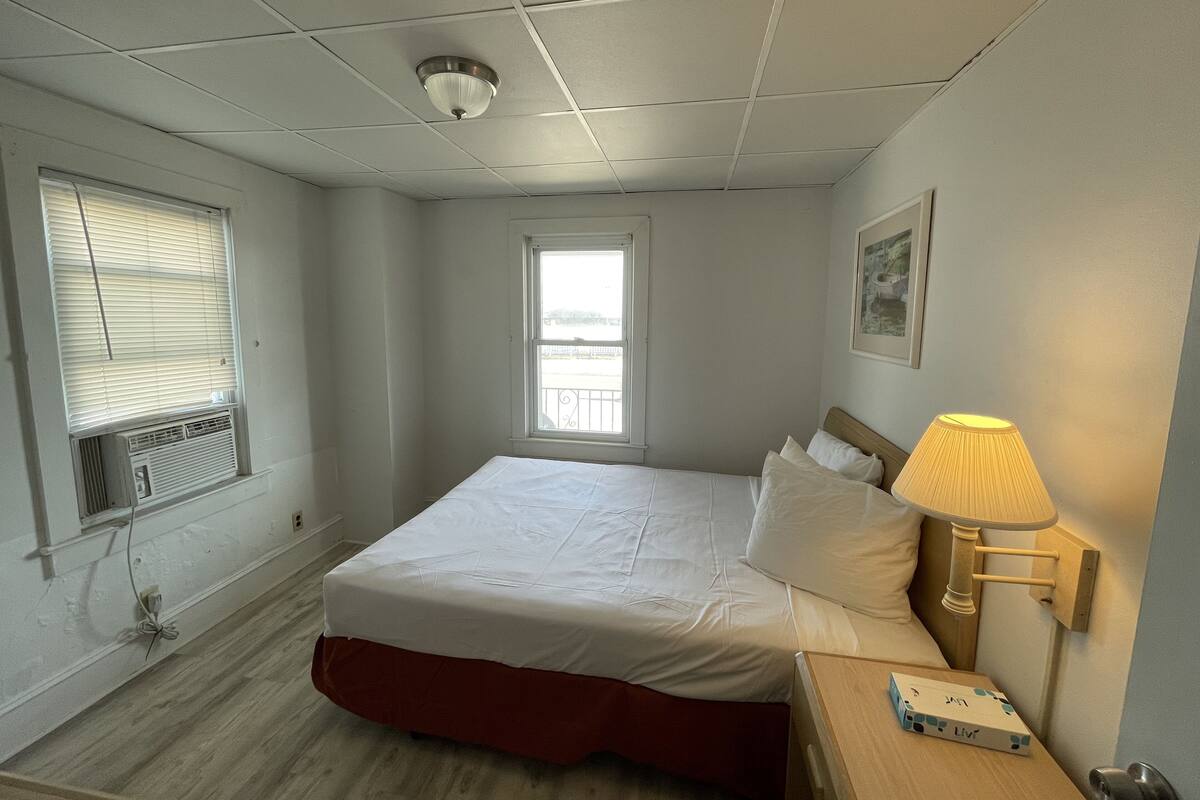 A hotel room with a bed, a bedside lamp, a window with blinds, and a ceiling light.