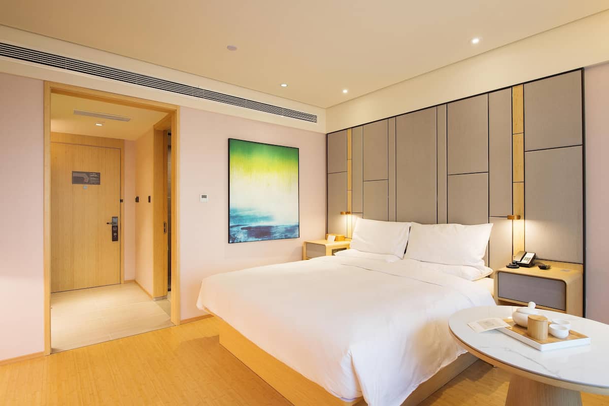 A hotel room with a large bed, bedside tables, a nightstand with a phone, a wall-mounted artwork, and a door leading to another room.