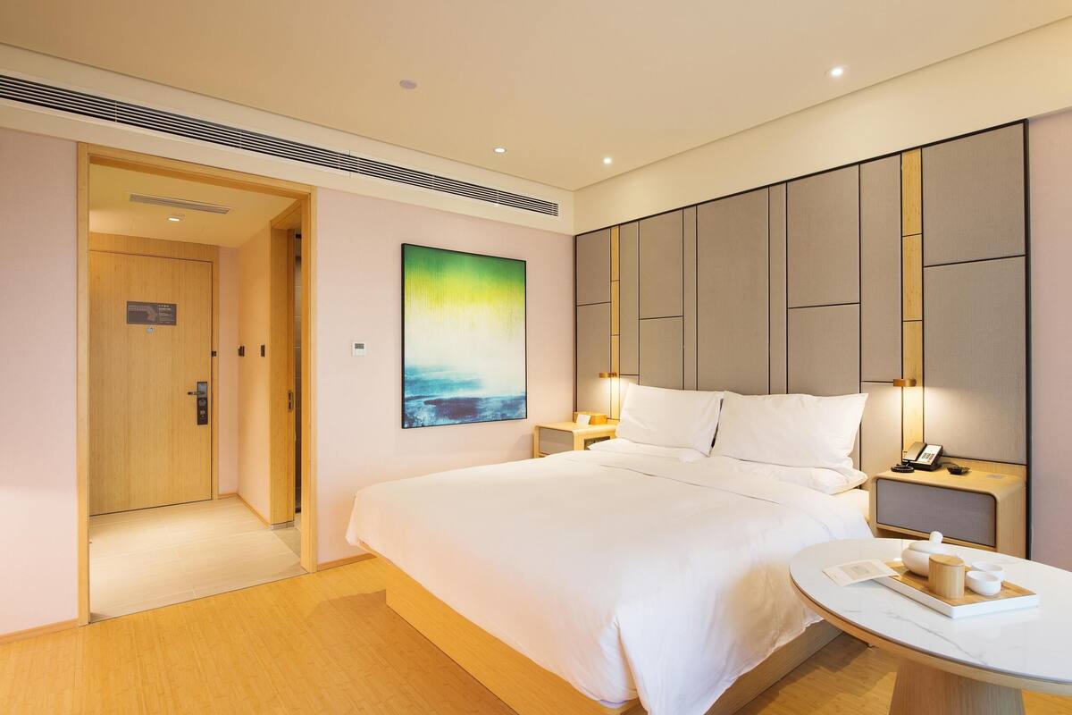 Una habitación de hotel con una cama grande, mesitas de noche, un buró con teléfono, una obra de arte en la pared y una puerta que da a otra habitación.