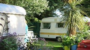 Uma caravana branca com uma faixa amarela estacionada em um jardim com diversas plantas e um carro vermelho.