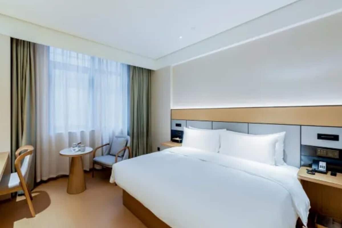 Une chambre d’hôtel avec un lit, une petite table, une chaise, un bureau et une fenêtre avec des rideaux.