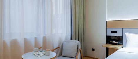 Quarto de hotel com cama, uma cadeira, uma mesinha com jogos de chá e uma mesa de cabeceira com abajur.