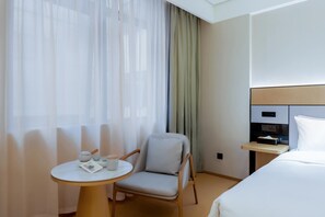 Hotellihuone, jossa on sänky, tuoli, pieni pöytä teekupposineen ja yöpöytä, jossa on lamppu.
