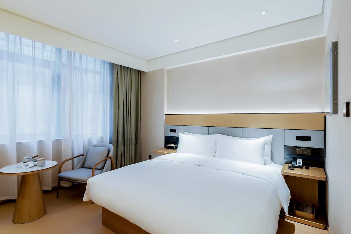 Une chambre d’hôtel avec un grand lit, une table de chevet, une chaise, une petite table et une fenêtre avec des rideaux.