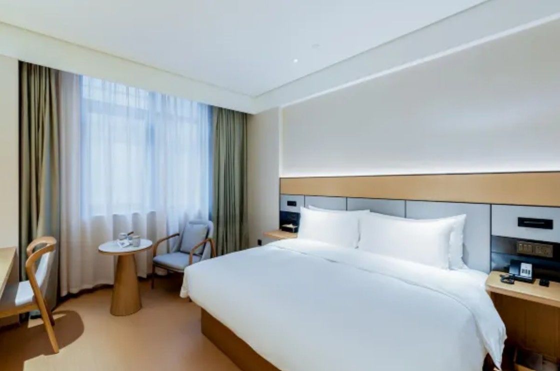 A hotel room with a bed, a small table, a chair, a desk, and a window with curtains.
