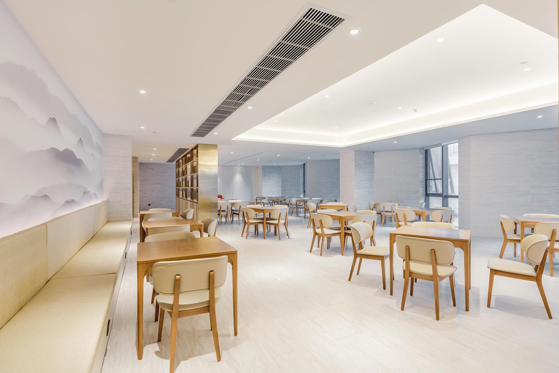 A modern dining area with wooden tables and chairs, a decorative wall with a mountain design, and a large window providing natural light.