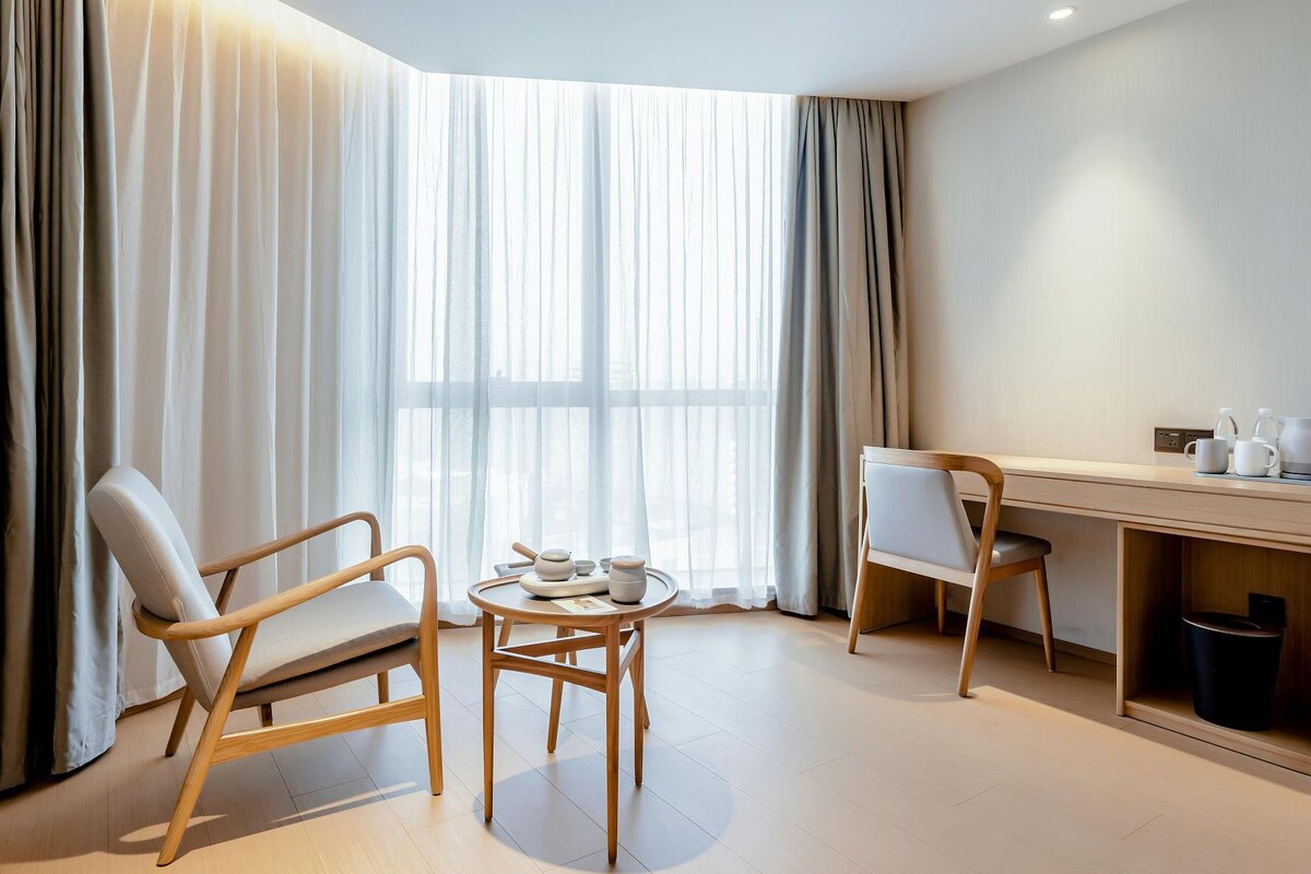 Moderni hotellihuone, jossa on puinen työpöytä, kaksi tuolia, pieni pyöreä pöytä teekupit ja -lautaset valmiina sekä läpikuultavat verhot.