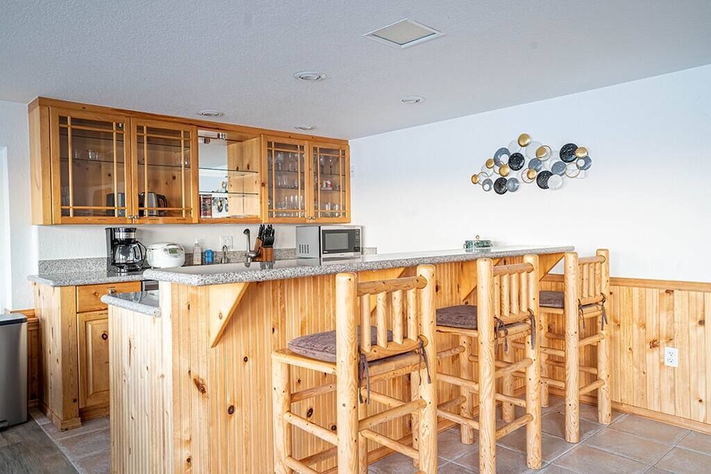 Eine Küche mit Holzausstattung, einer Arbeitsplatte aus Granit, einer Mikrowelle und einer Kaffeemaschine. Es gibt drei hölzerne Barhocker und eine an der Wand befestigte Tellerablage.