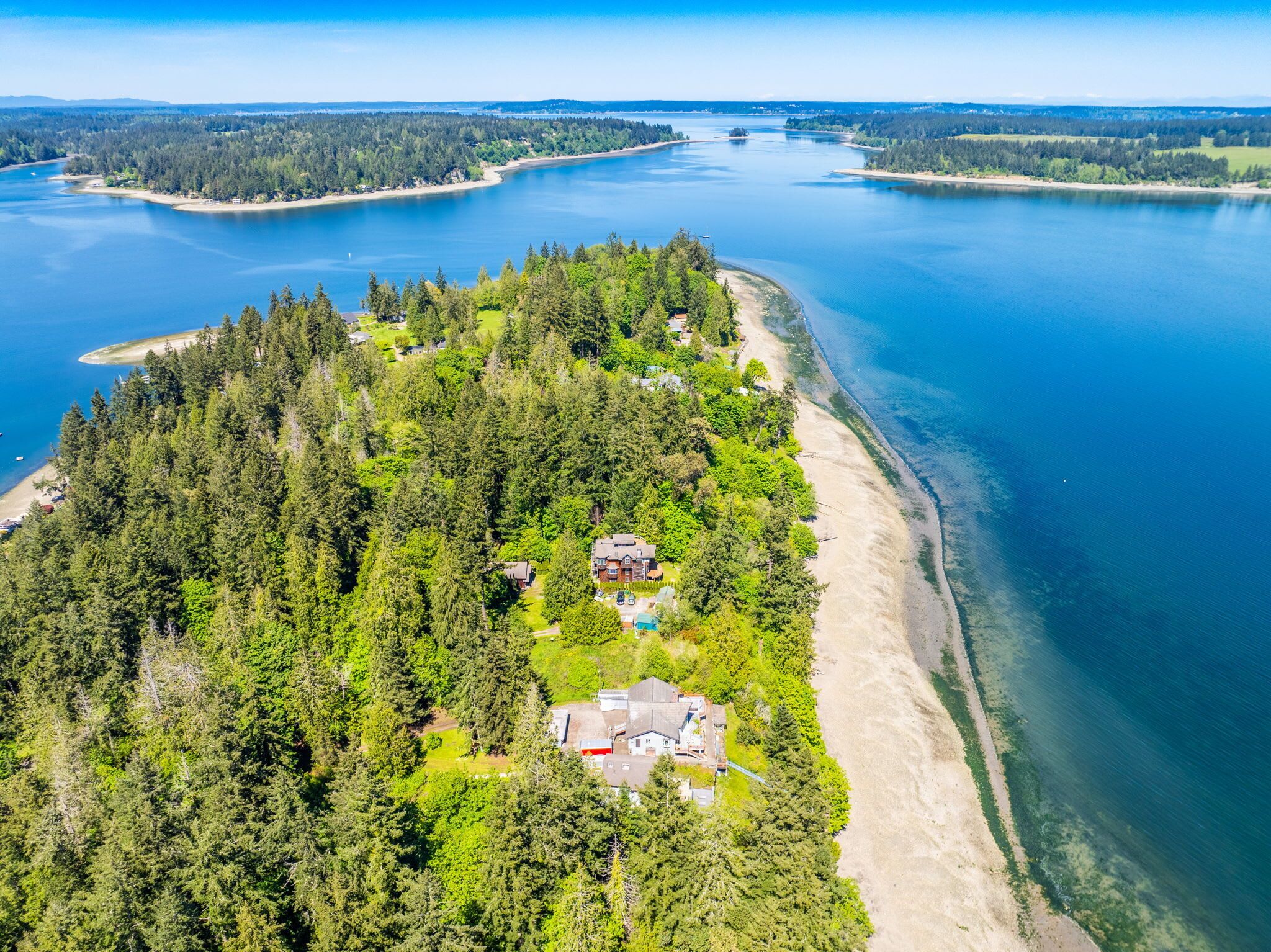 An aerial view of a coastal property with a large body of water, dense forest, and a sandy beach.