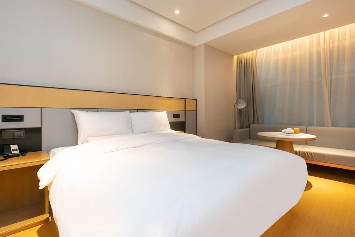 A modern hotel room with a large bed, a bedside table with a phone, a small round table with a lamp, and a sofa.