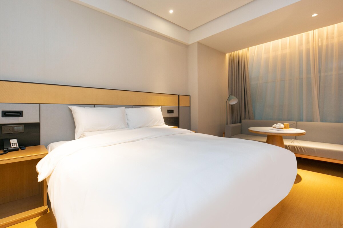 A modern hotel room with a large bed, a bedside table with a phone, a small round table with a lamp, and a sofa.