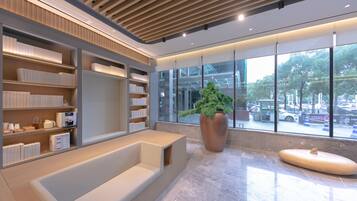 A modern lobby with a seating area, a large potted plant, and a view of the street through large windows.