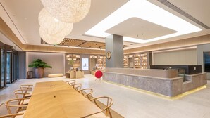 En moderne hotellobby med en lang træreception, en stor stueplante og et siddeområde med træstole.