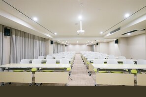 Un salón de conferencias con filas de mesas y sillas, una mesa grande al frente y una pantalla de proyector.