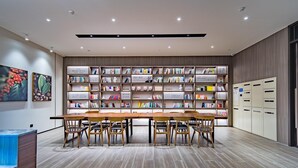 A modern library with a long wooden table and chairs, surrounded by bookshelves filled with books.