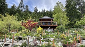 A wooden cabin with a balcony, surrounded by a garden with various plants and rocks.
