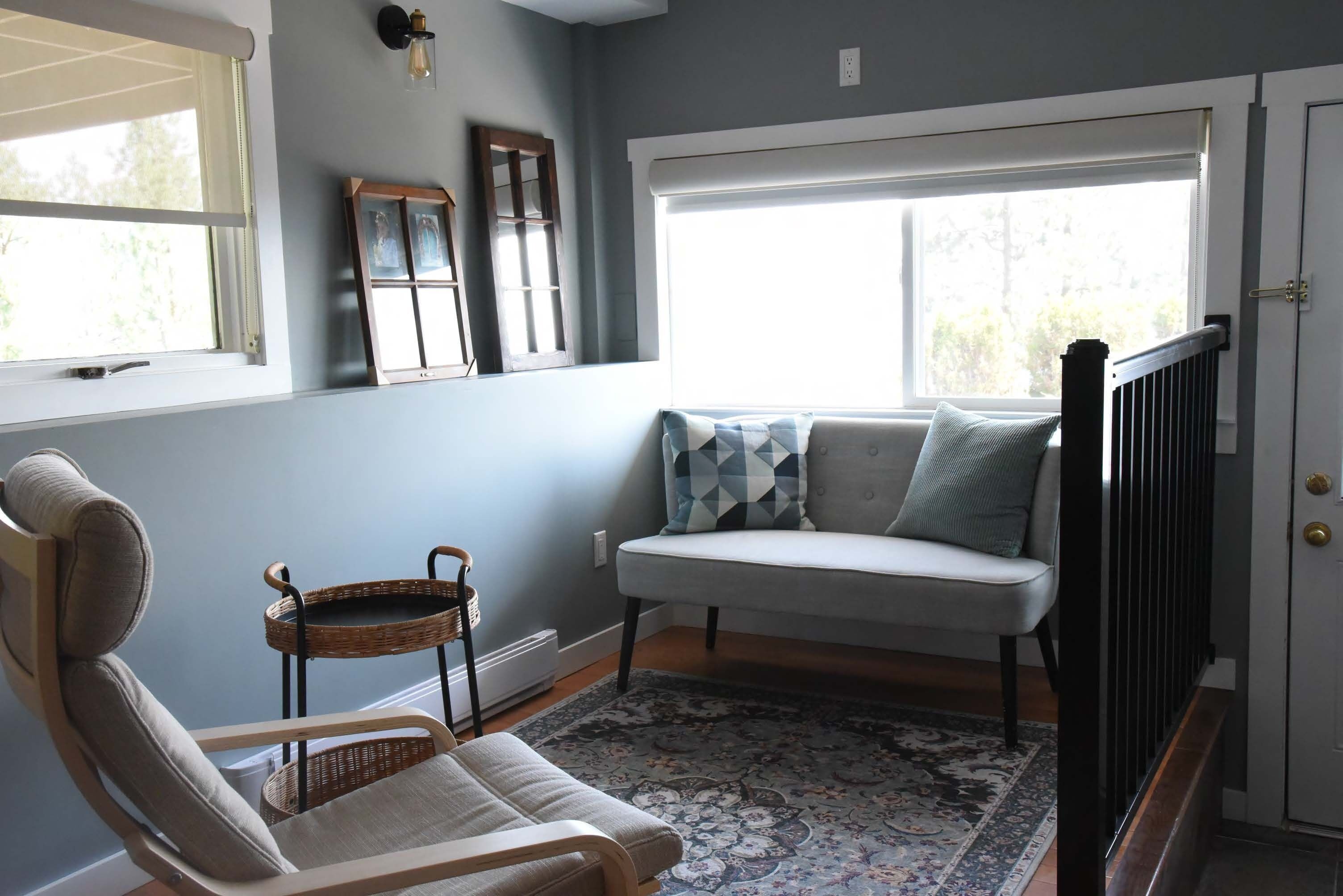 A room with a light blue wall, a white sofa with patterned cushions, a wicker chair, and a patterned rug.