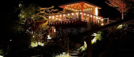 A wooden gazebo with string lights, surrounded by lush greenery and a stone staircase leading up to it.