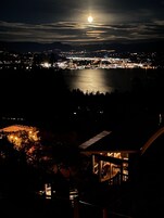 Vista nocturna de una ciudad costera con una luna destacada en el cielo, un puerto iluminado y un edificio con un balcón visible.