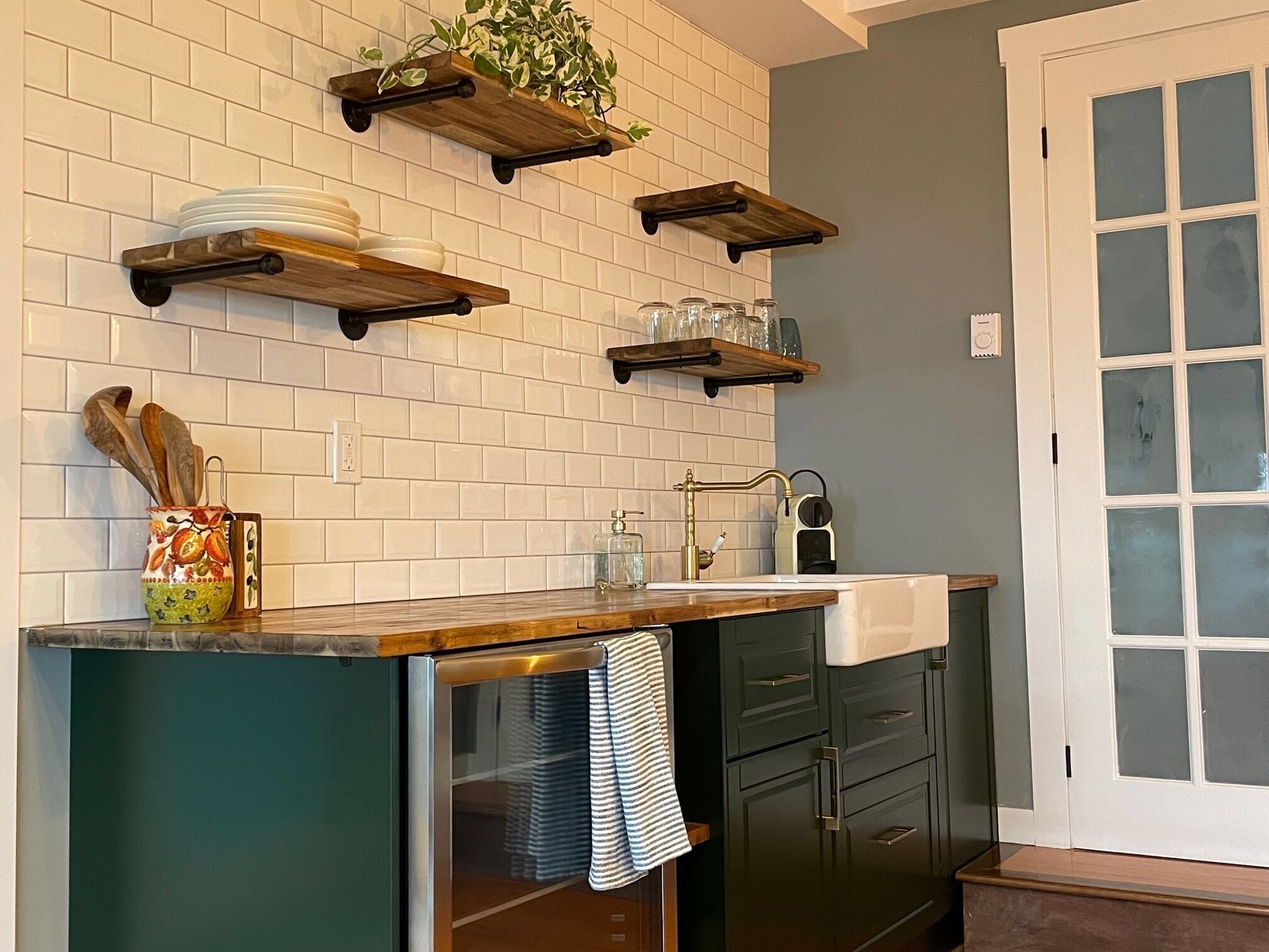 Una cocina con azulejos blancos tipo metro, estantes de madera, un refrigerador de acero inoxidable y un ventanal con vidrio esmerilado.