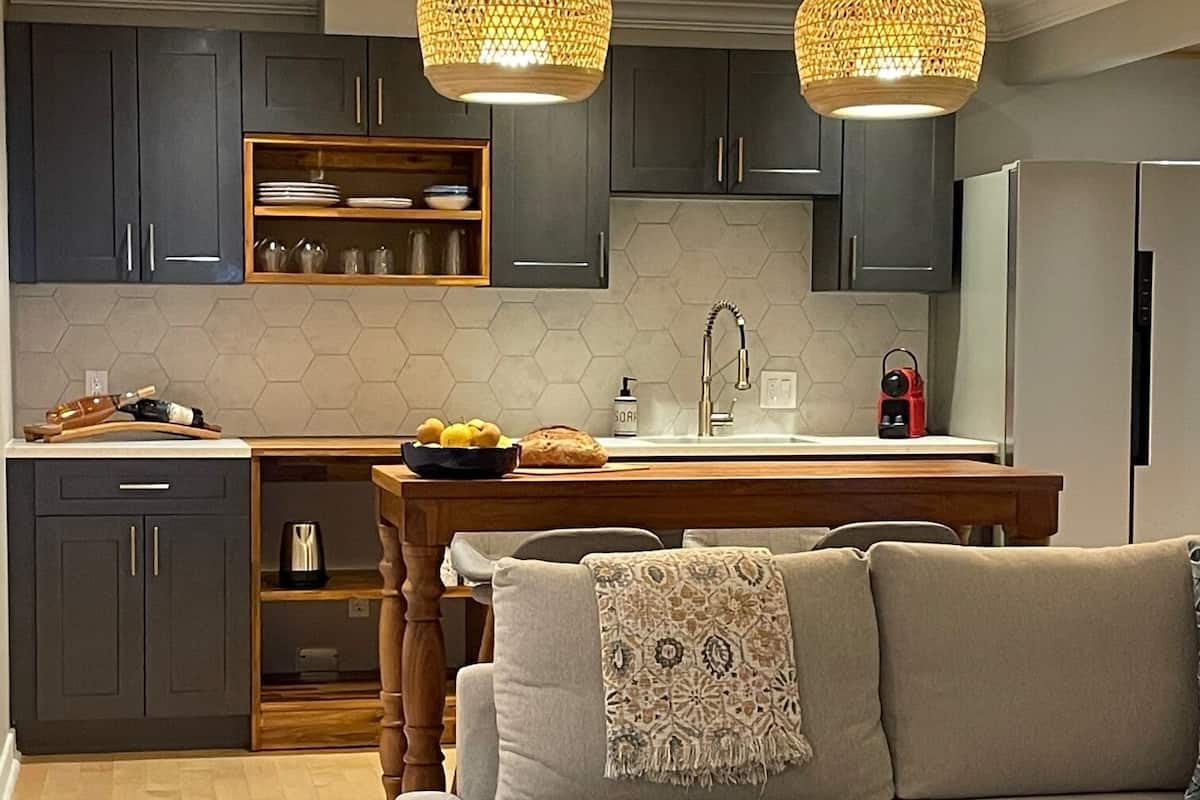 A modern kitchen with dark cabinets, a wooden dining table, and a built-in oven. There is a sofa with a patterned throw blanket and a wicker chair.