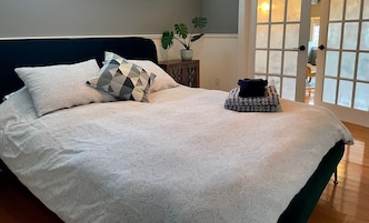 Una cama bien hecha con ropa de cama estampada, un cojín geométrico y una manta doblada sobre un piso de madera.