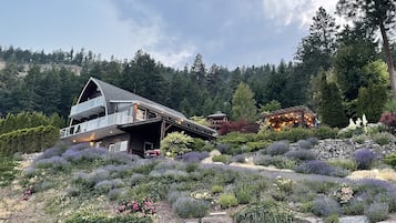 Una casa moderna de dos pisos con techo inclinado y balcón, rodeada de un jardín ajardinado con diversas plantas y un muro de piedra.