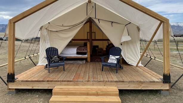 A wooden tent with a pitched roof, two blue Adirondack chairs, and a bed inside. - Yellowstone Dreamin Camp (Pray)