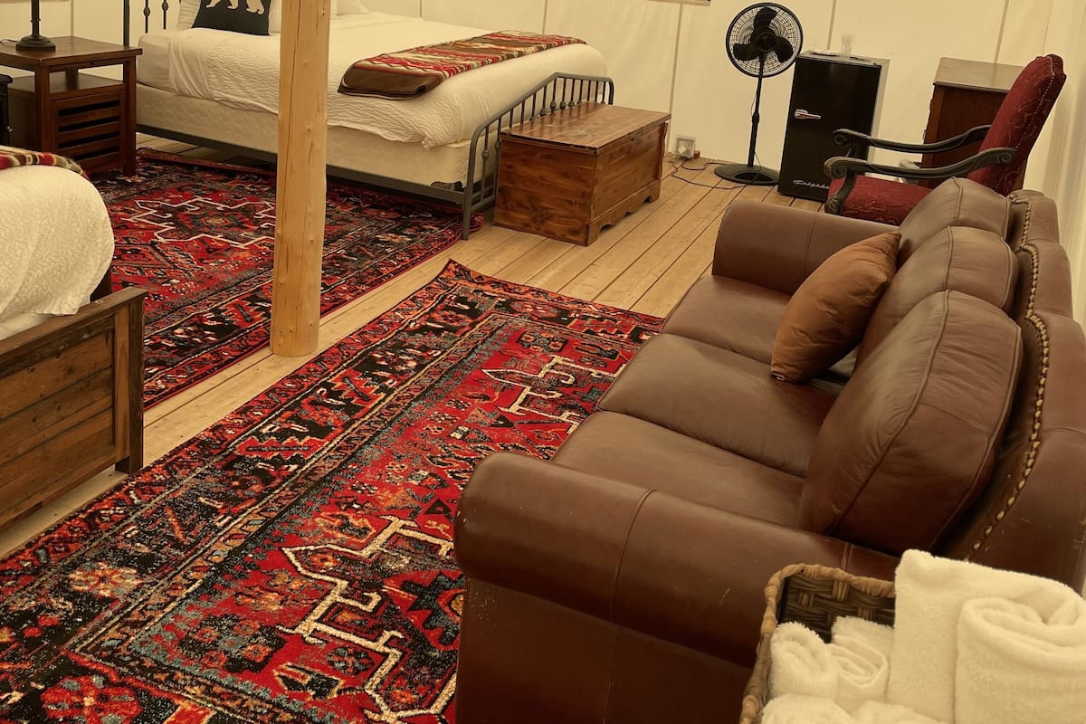 A tent-style room with a bed, a wooden table, a brown leather sofa, a red and black patterned rug, and a basket of white towels.