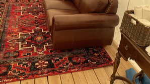 A brown leather sofa with a patterned rug, a wooden floor, a red chair, a fan, and a small wooden cabinet. - Yellowstone Dreamin Camp (Pray)