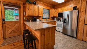 A wooden cabin kitchen with a central island, stainless steel appliances, and a view of a cabin through the window. - Groot's Place - 5min from parkway - massage chair/pool table/foosball (Pigeon Forge)
