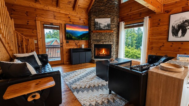 A log cabin interior with a stone fireplace, black leather furniture, and a view of the outdoors. - Groot's Place - 5min from parkway - massage chair/pool table/foosball (Pigeon Forge)