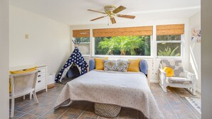 Ein Schlafzimmer mit Bett, Deckenventilator, Fenster mit Blick auf Grünflächen, einem weißen Stuhl und einem kleinen weißen Tisch.