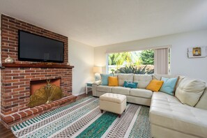 Un salon doté d’une cheminée en briques, d’un téléviseur à écran plat fixé au-dessus, d’un canapé beige agrémenté de coussins colorés et d’un tapis à motifs.