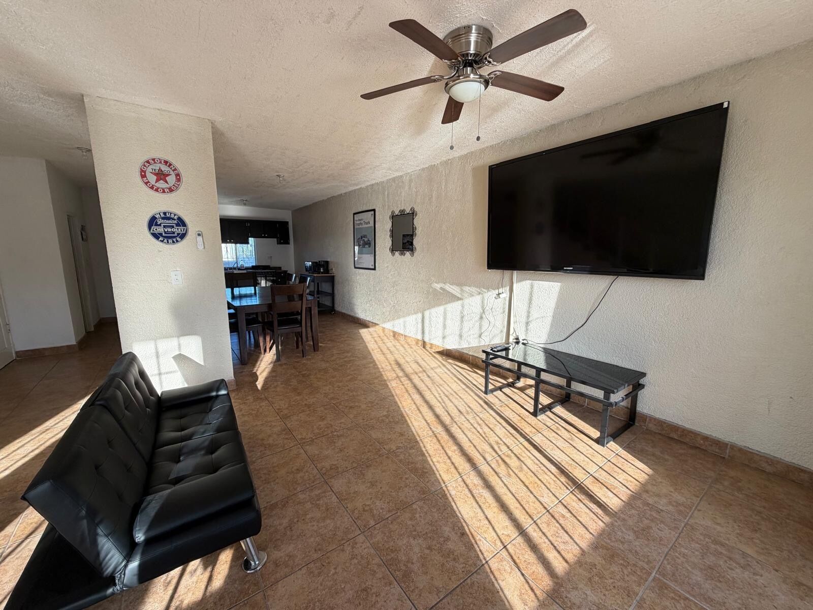 A spacious living room with a flat-screen TV mounted on the wall, a ceiling fan, and a black leather couch.