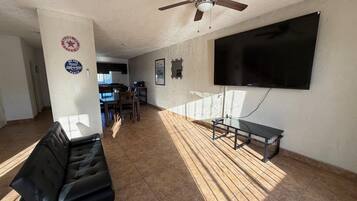 A spacious living room with a flat-screen TV mounted on the wall, a ceiling fan, and a black leather couch.