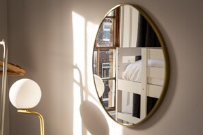 A room with a round mirror reflecting a bed and a window with daylight.