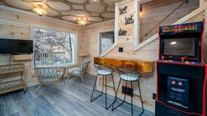 Interior de una cabaña de madera con una zona de bar, dos taburetes, un televisor y una máquina arcade de Mortal Kombat.